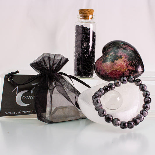Snowflake obsidian bracelet, rhodonite heart-shaped stone, and jar with black tourmaline stone chips. Selenite triangular bowl with mini crystal quartz sphere.